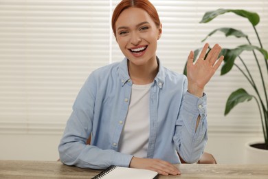 Young woman waving hello during video chat at wooden table indoors, view from web camera