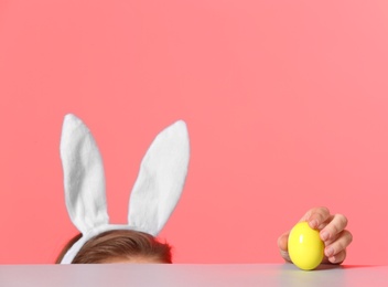 Little girl wearing bunny ears headband  and playing with Easter egg at table against color background