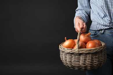 Photo of Woman holding wicker basket with raw yellow onion bulbs on black background, closeup. Space for text