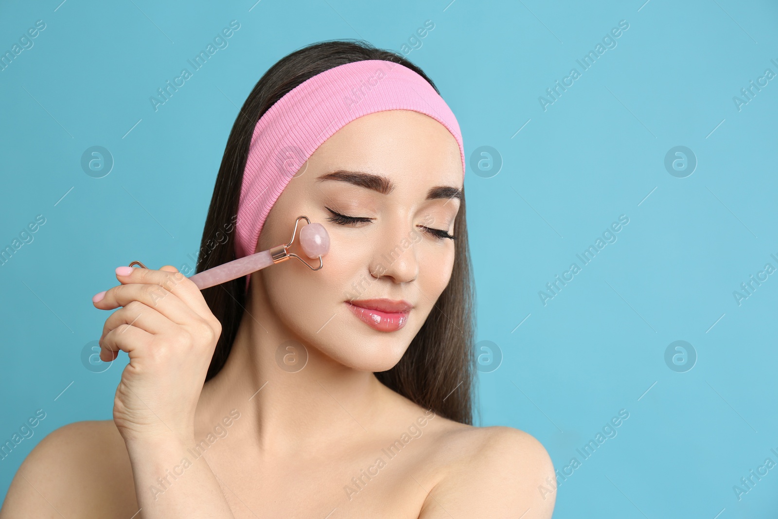 Photo of Woman using natural pink quartz face roller on light blue background