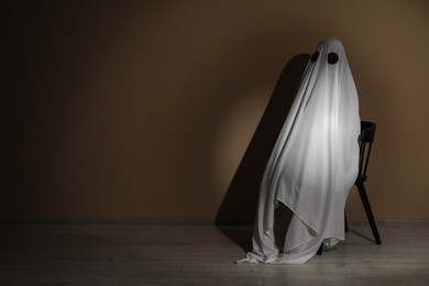 Photo of Creepy ghost. Woman covered with sheet sitting on chair near brown wall, space for text