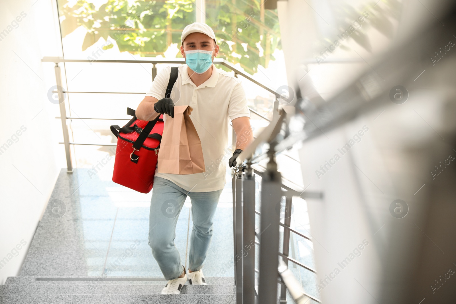 Photo of Courier in protective mask and gloves with order indoors. Restaurant delivery service during coronavirus quarantine