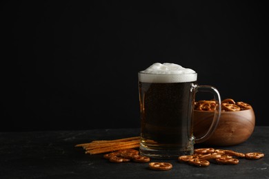 Photo of Delicious pretzel crackers, glass of beer and salt sticks on black table against dark background, space for text