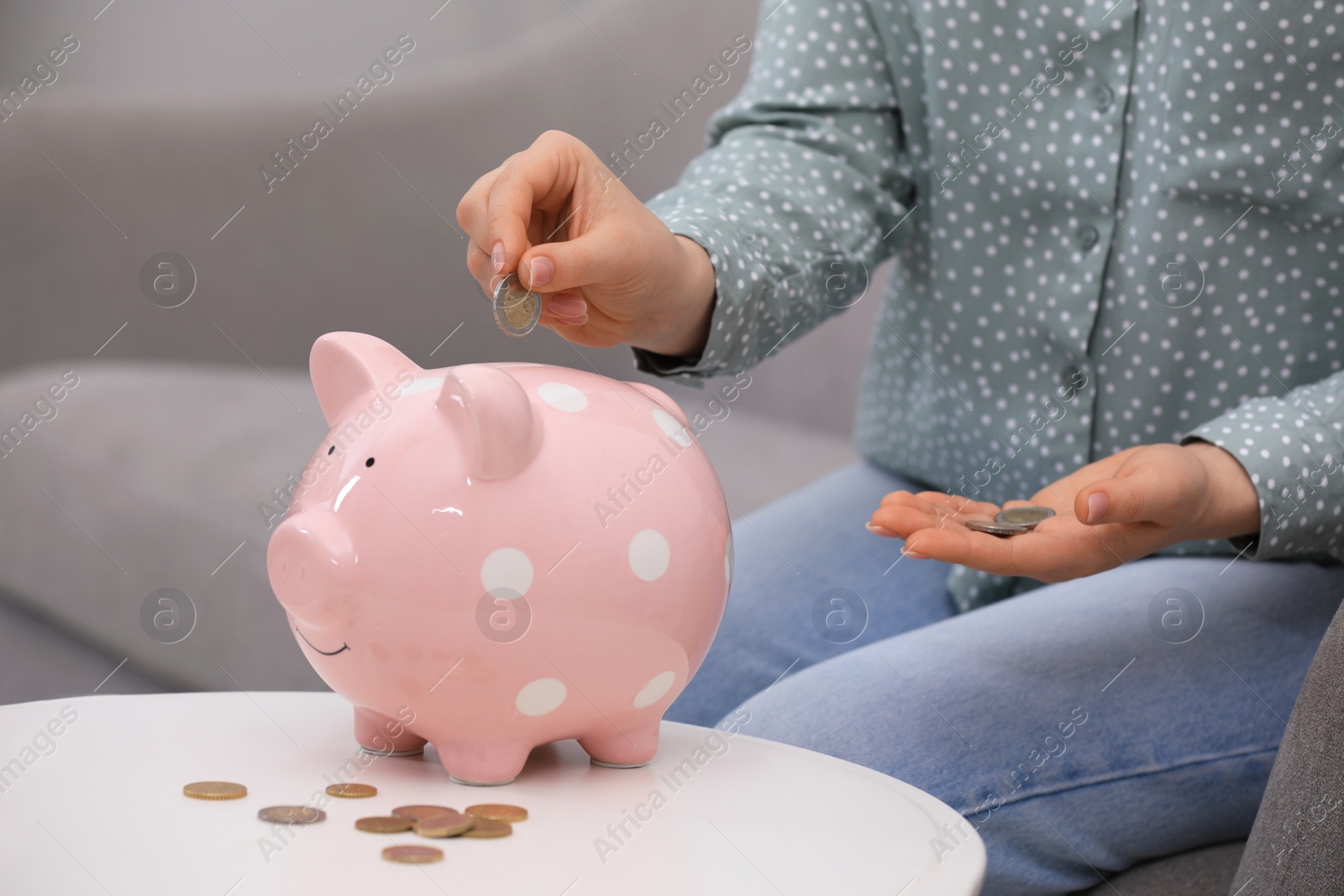 Photo of Young woman putting coin into piggy bank at table indoors, closeup