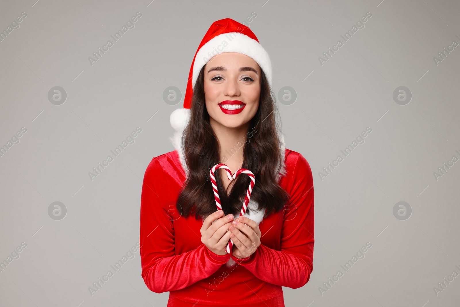 Photo of Christmas celebration. Beautiful young woman in Santa hat and red dress with candy canes on grey background
