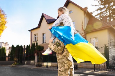 Photo of Soldier in military uniform carrying his daughter on shoulders and Ukrainian flag on city street, space for text. Family reunion