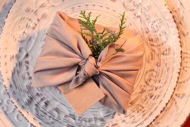 Photo of Furoshiki technique. Gift packed in beige fabric with blank card on plate, top view