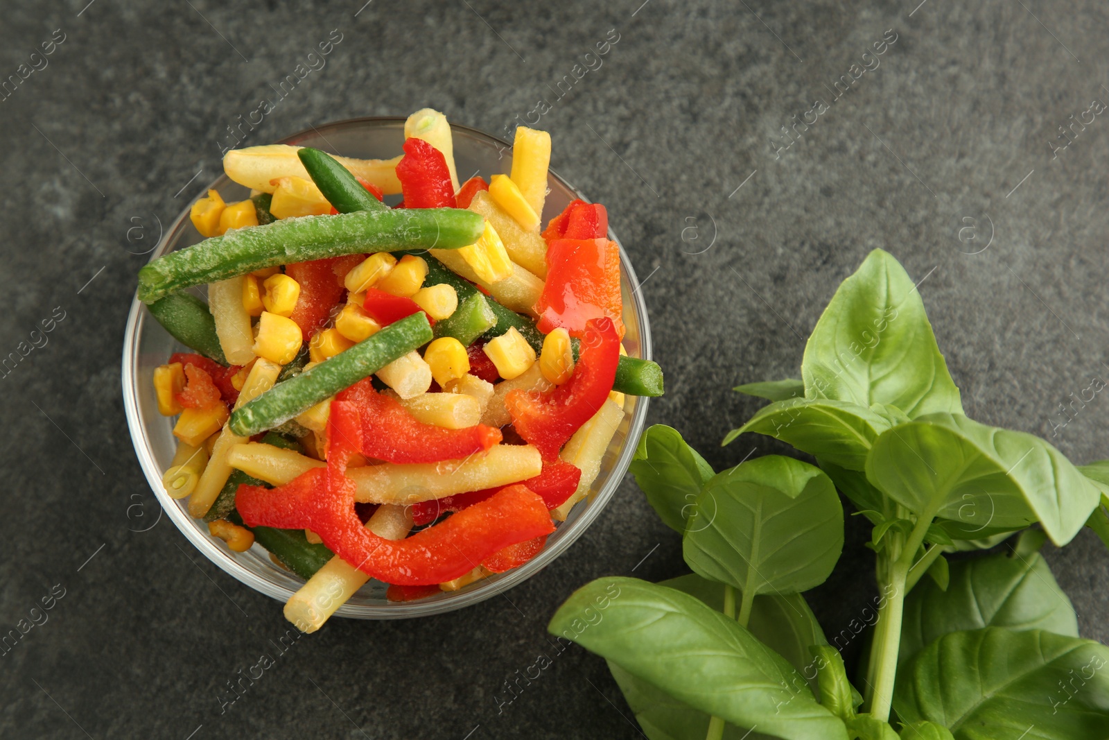 Photo of Bowl of different frozen vegetables and fresh basil on grey table, top view