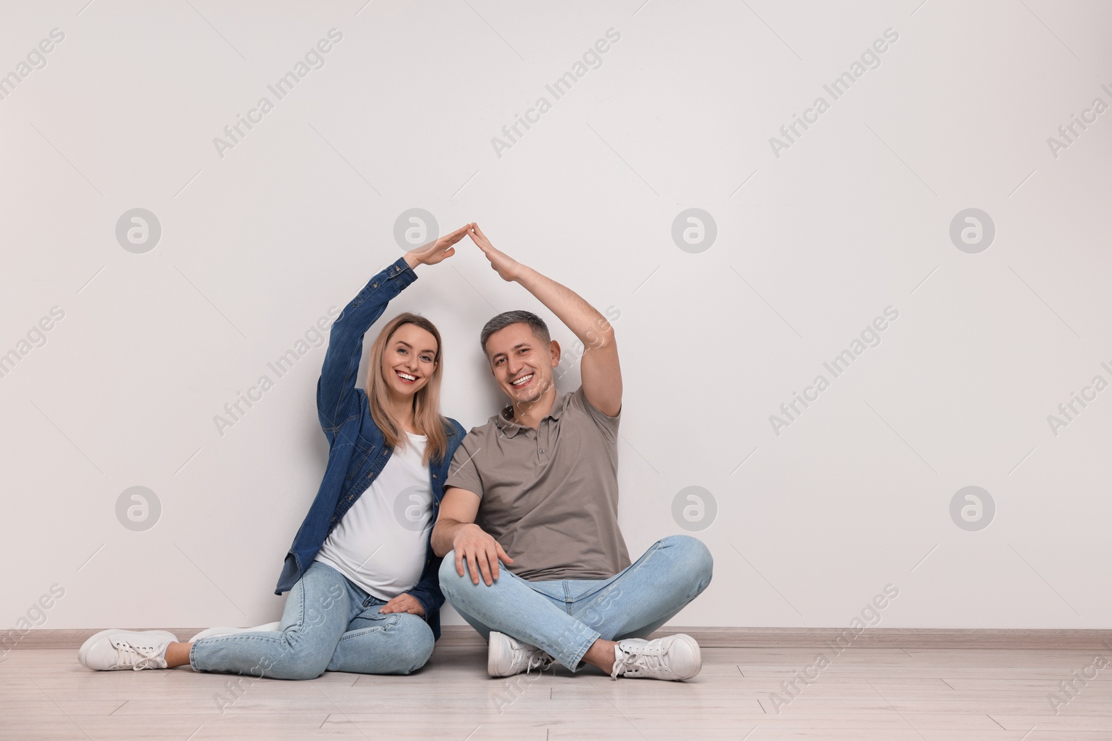 Photo of Pregnant woman with her husband forming roof with their hands while sitting on floor indoors. Space for text