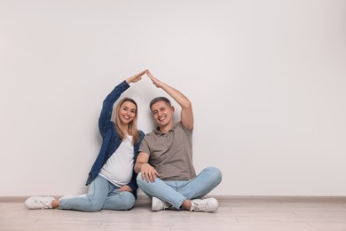Pregnant woman with her husband forming roof with their hands while sitting on floor indoors. Space for text