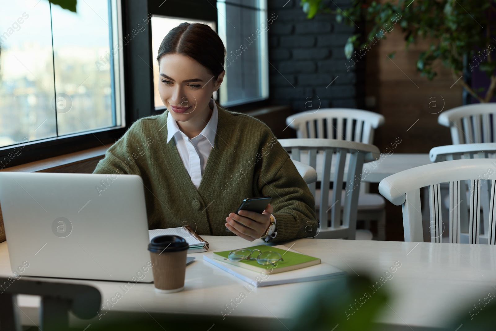 Photo of Young female student with laptop using smartphone while studying at table in cafe