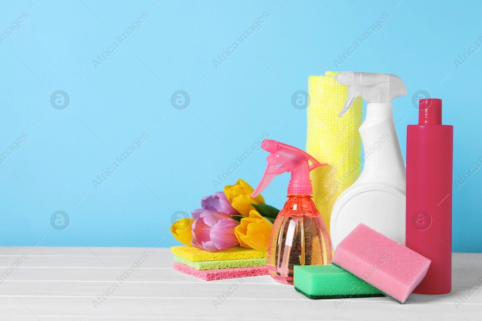 Photo of Spring cleaning. Detergents, flowers and sponges on white wooden table against light blue background. Space for text