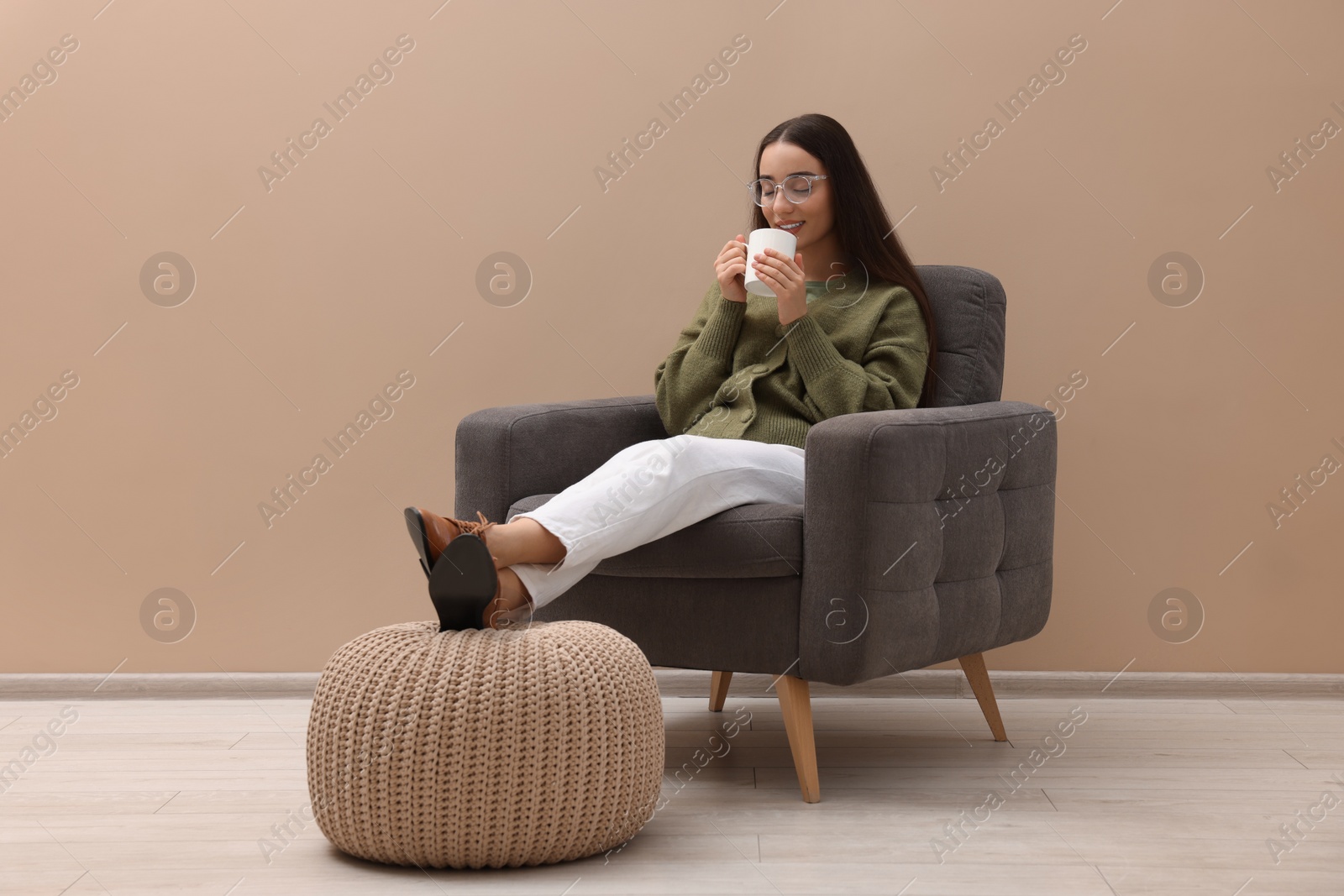 Photo of Beautiful woman with cup of drink sitting in armchair near beige wall indoors