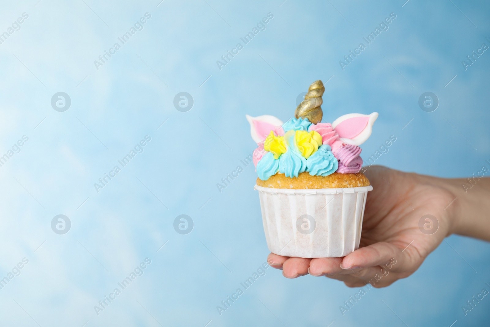 Photo of Woman holding cute sweet unicorn cupcake on light blue background, closeup. Space for text