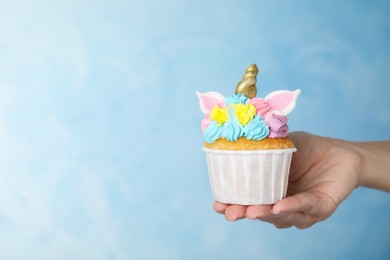 Photo of Woman holding cute sweet unicorn cupcake on light blue background, closeup. Space for text