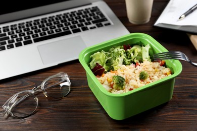 Photo of Container with tasty food, fork, laptop and glasses on wooden table. Business lunch