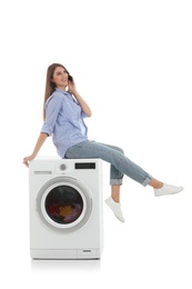 Young woman with phone sitting on washing machine against white background. Laundry day