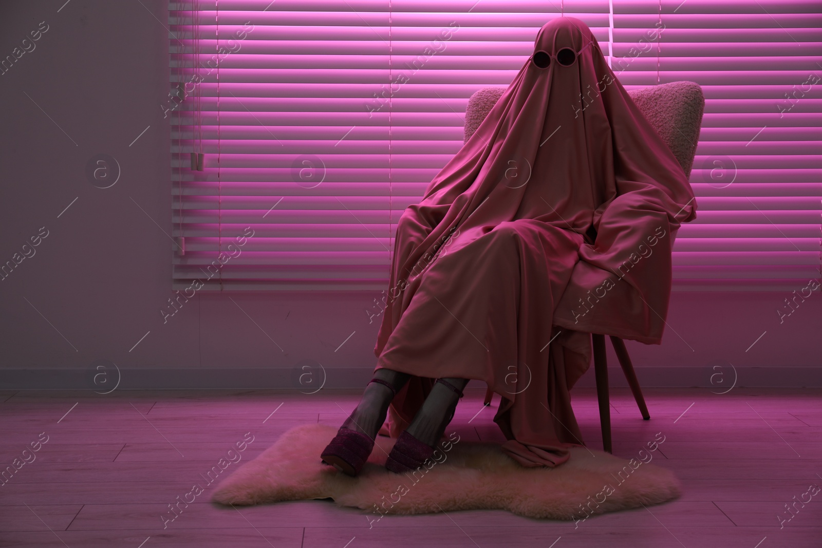 Photo of Glamorous ghost. Woman in sheet and high heel shoes on armchair in pink light, space for text