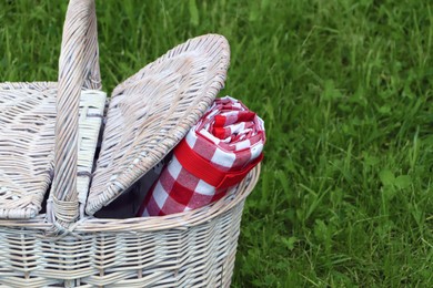 Photo of Rolled checkered tablecloth in picnic basket on green grass outdoors, closeup. Space for text