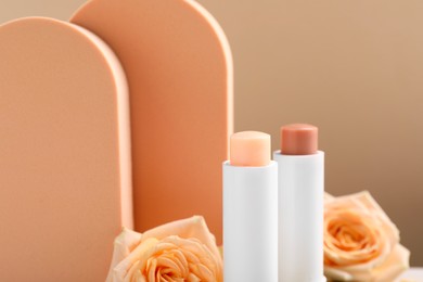 Photo of Different lip balms and rose flowers against light brown background