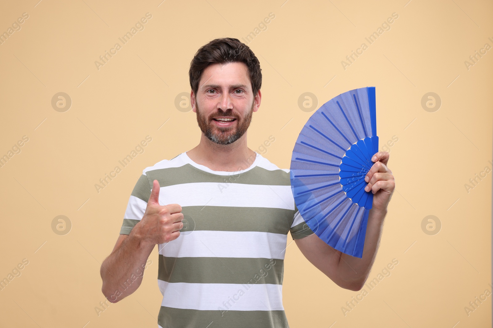 Photo of Happy man holding hand fan and showing thumb up on beige background