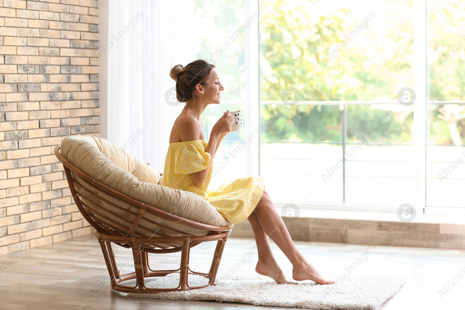 Photo of Young woman with cup of aromatic coffee sitting in papasan chair near window at home