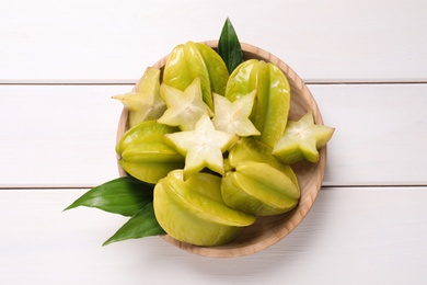 Photo of Cut and whole carambolas with green leaves in bowl on white wooden table, top view