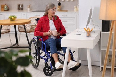 Woman in wheelchair with cup of drink using computer at home