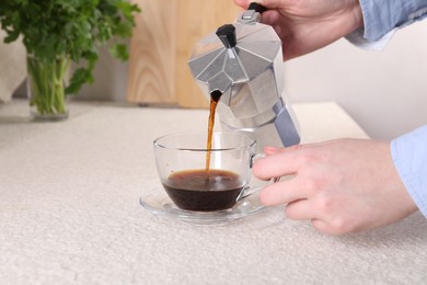 Woman pouring aromatic coffee from moka pot into glass cup at light textured table, closeup. Space for text