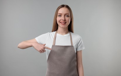 Photo of Beautiful young woman pointing at kitchen apron on grey background. Mockup for design