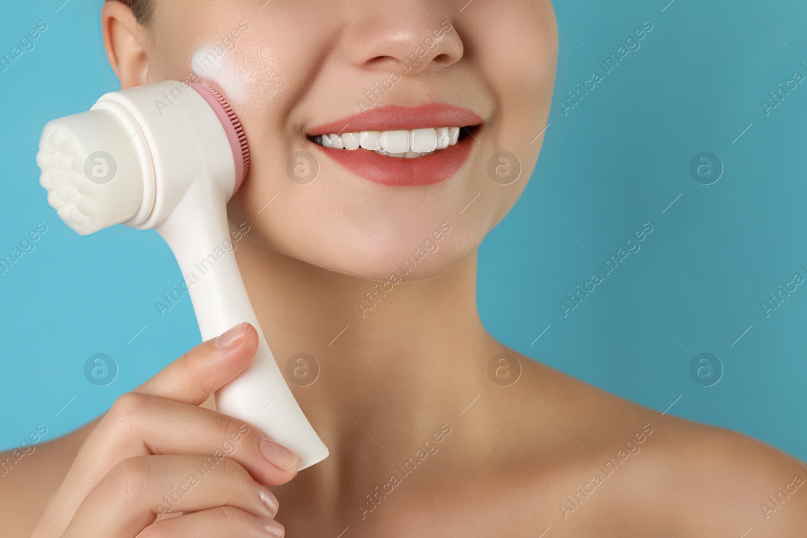 Photo of Young woman washing face with brush and cleansing foam on light blue background, closeup. Cosmetic products