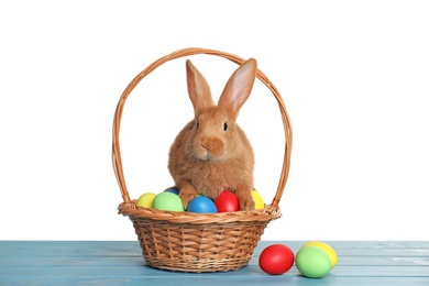 Photo of Cute fluffy bunny and wicker basket with Easter eggs on light blue wooden table