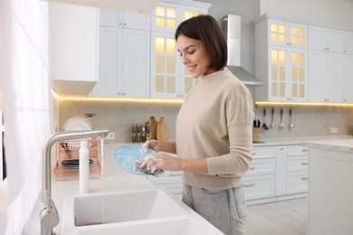 Photo of Happy young woman washing plate above sink in modern kitchen
