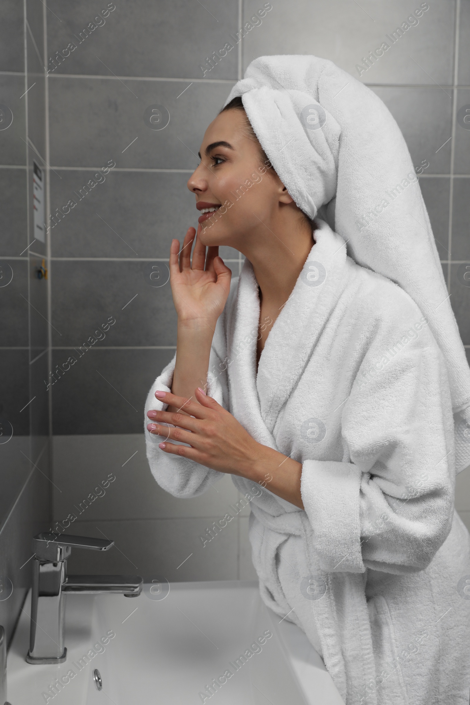 Photo of Beautiful young woman in bathrobe with towel on head near mirror at home