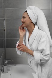 Photo of Beautiful young woman in bathrobe with towel on head near mirror at home
