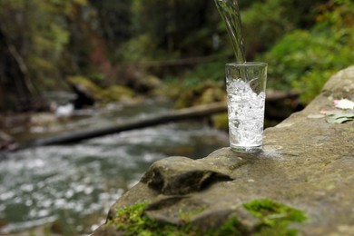 Photo of Fresh water pouring into glass on stone near stream in forest. Space for text