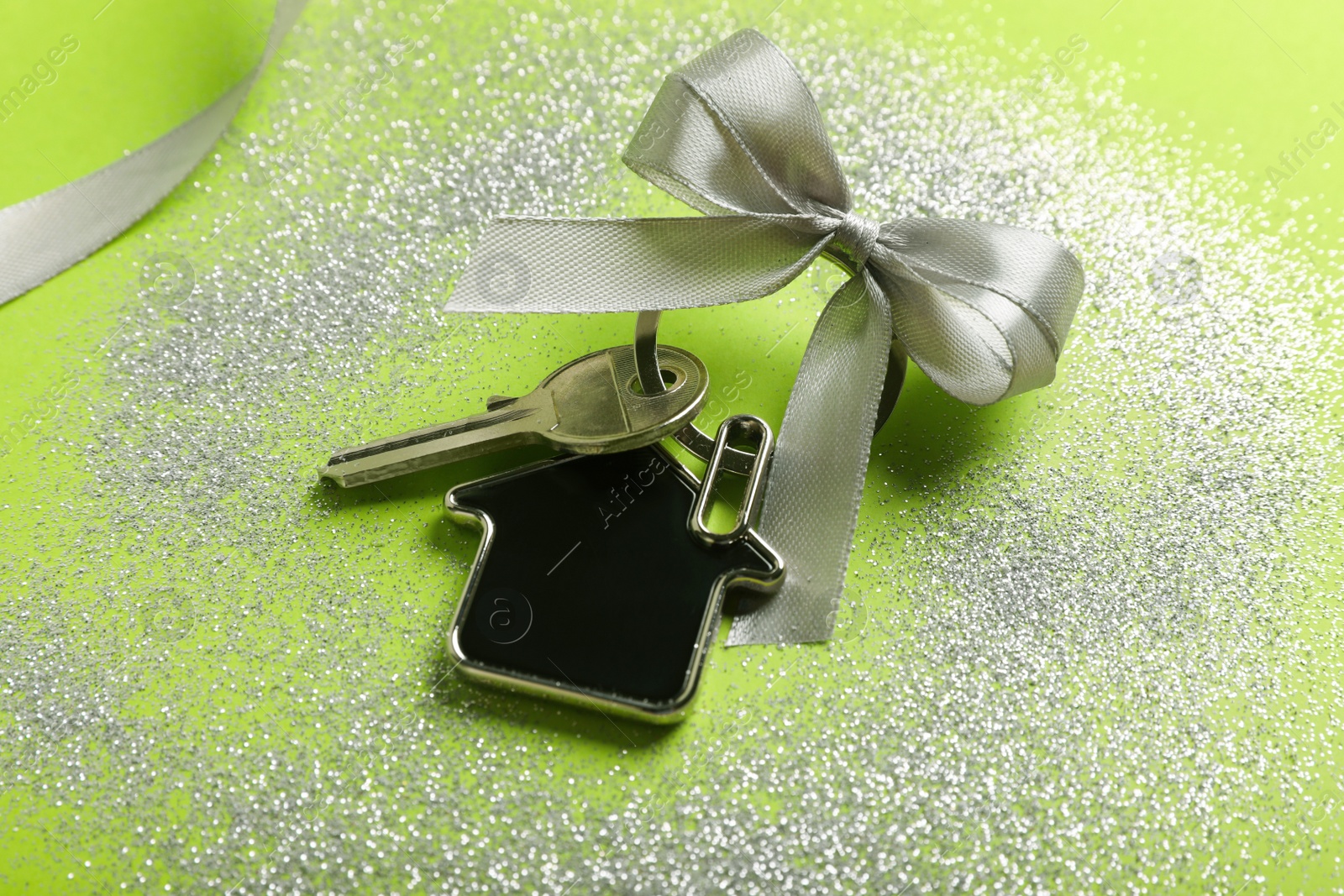 Photo of Key with trinket in shape of house, bow, ribbon and glitter on light green background, closeup. Housewarming party