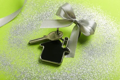 Key with trinket in shape of house, bow, ribbon and glitter on light green background, closeup. Housewarming party
