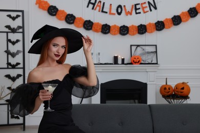Photo of Young woman in scary witch costume with glass of cocktail indoors, space for text. Halloween celebration