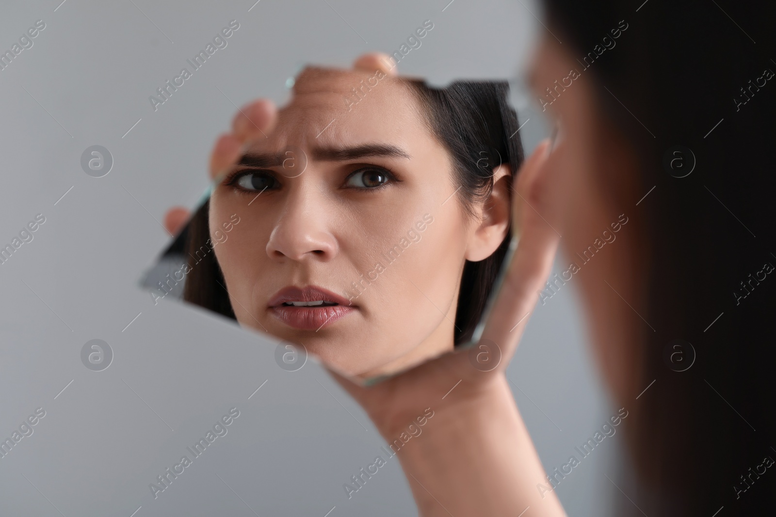 Photo of Young woman looking at herself in shard of broken mirror on light grey background, closeup