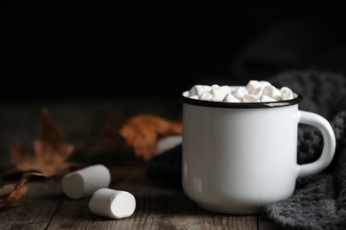 Photo of Mug of hot cozy drink with marshmallows, sweater and autumn leaves on table. Space for text