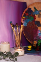 Photo of Brushes with colorful paints, wooden artist's palette and burning scented candles on light gray table