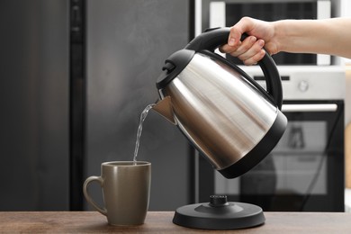 Photo of Woman pouring hot water from electric kettle into cup in kitchen, closeup