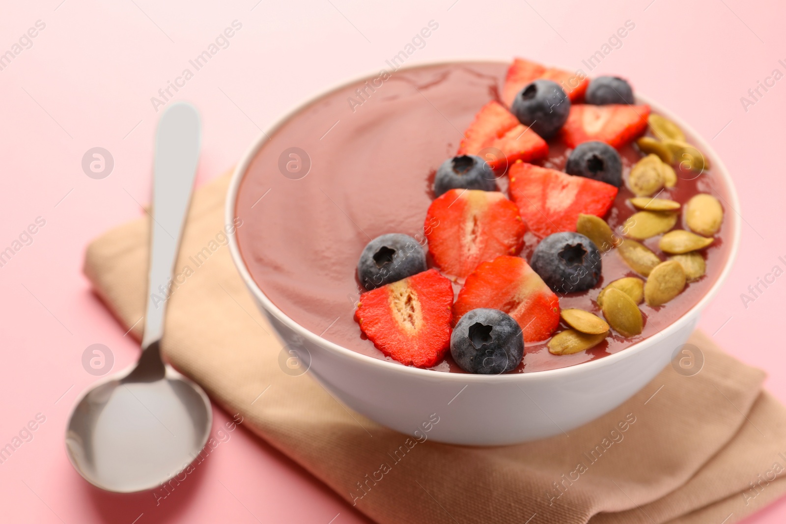 Photo of Bowl of delicious smoothie with fresh blueberries, strawberries and pumpkin seeds on pale pink background, closeup