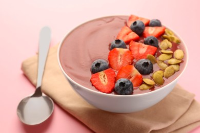 Bowl of delicious smoothie with fresh blueberries, strawberries and pumpkin seeds on pale pink background, closeup