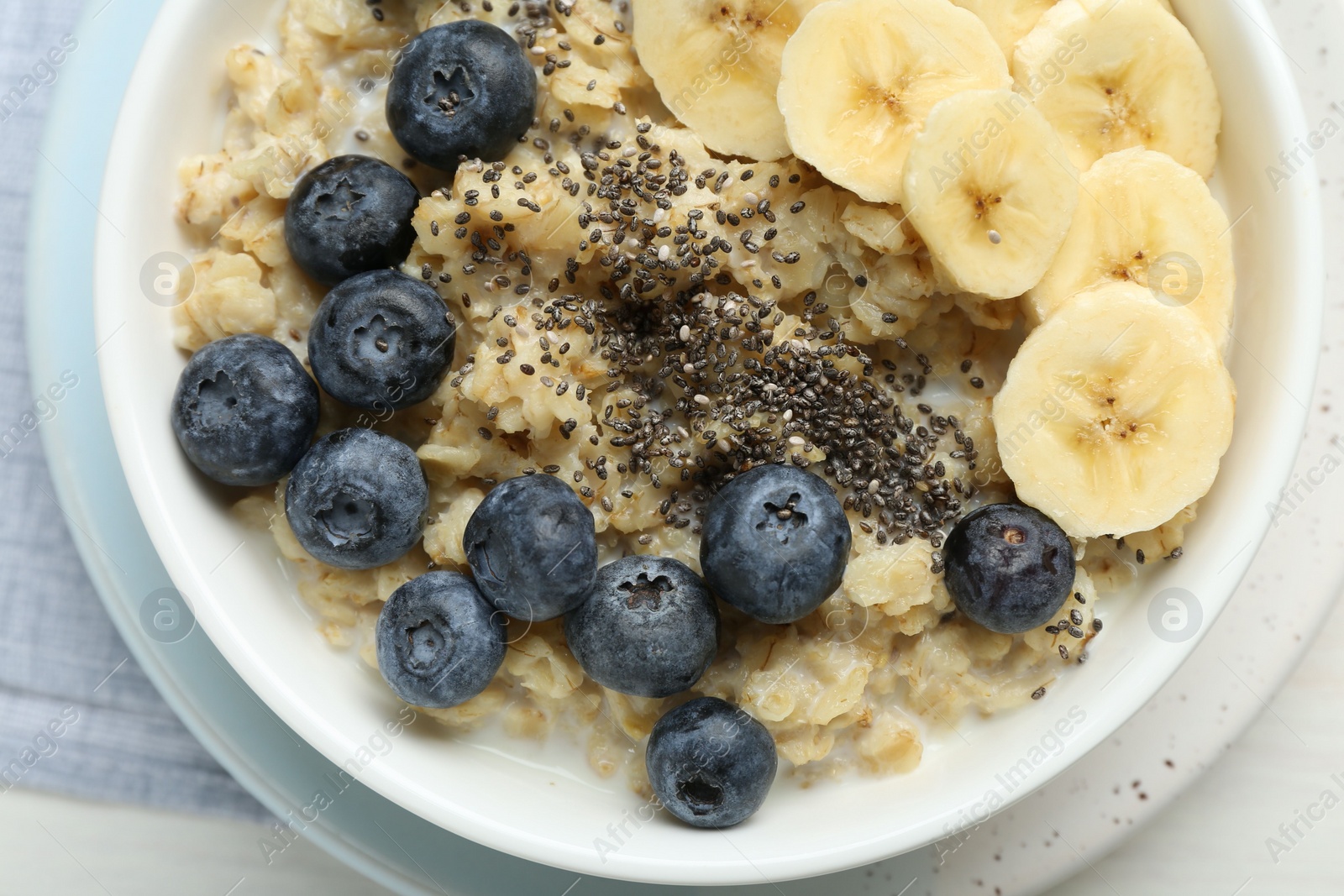 Photo of Tasty oatmeal with banana, blueberries and chia seeds served in bowl on white wooden table, top view