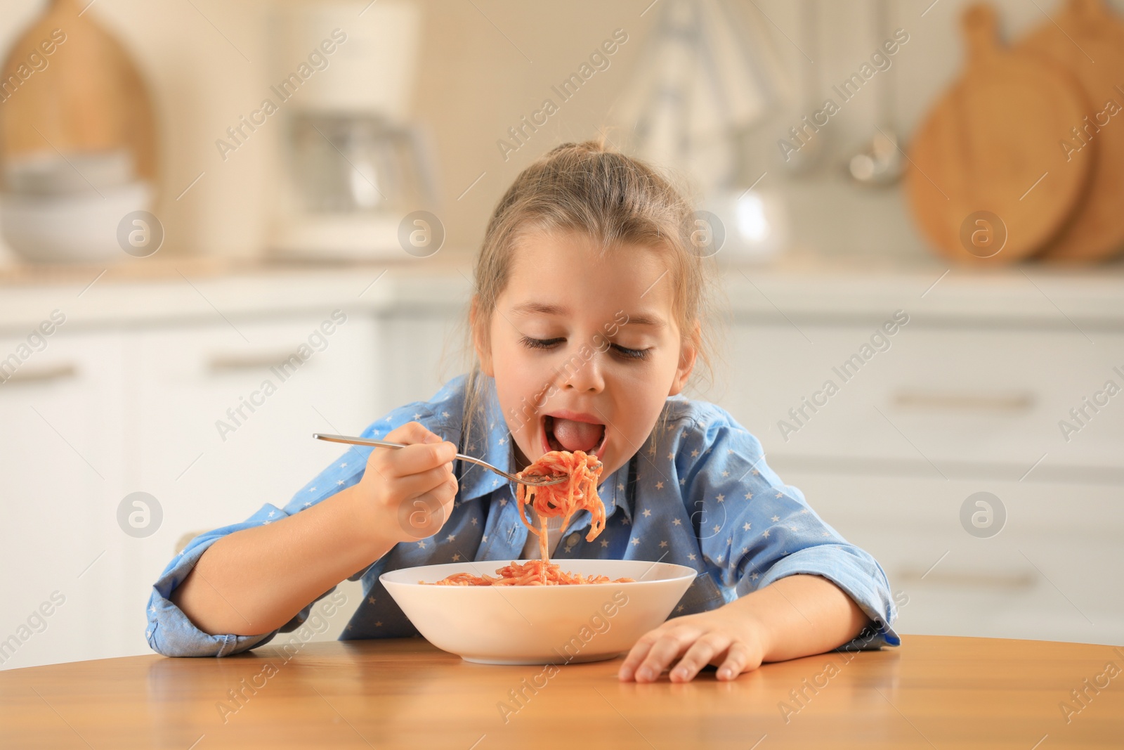 Photo of Cute little girl eating tasty pasta at table in kitchen