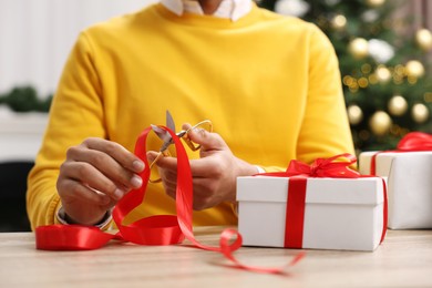 Photo of Christmas gift. Man cutting red ribbon at wooden table indoors, closeup