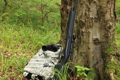 Hunting rifle, backpack and binoculars near tree in forest
