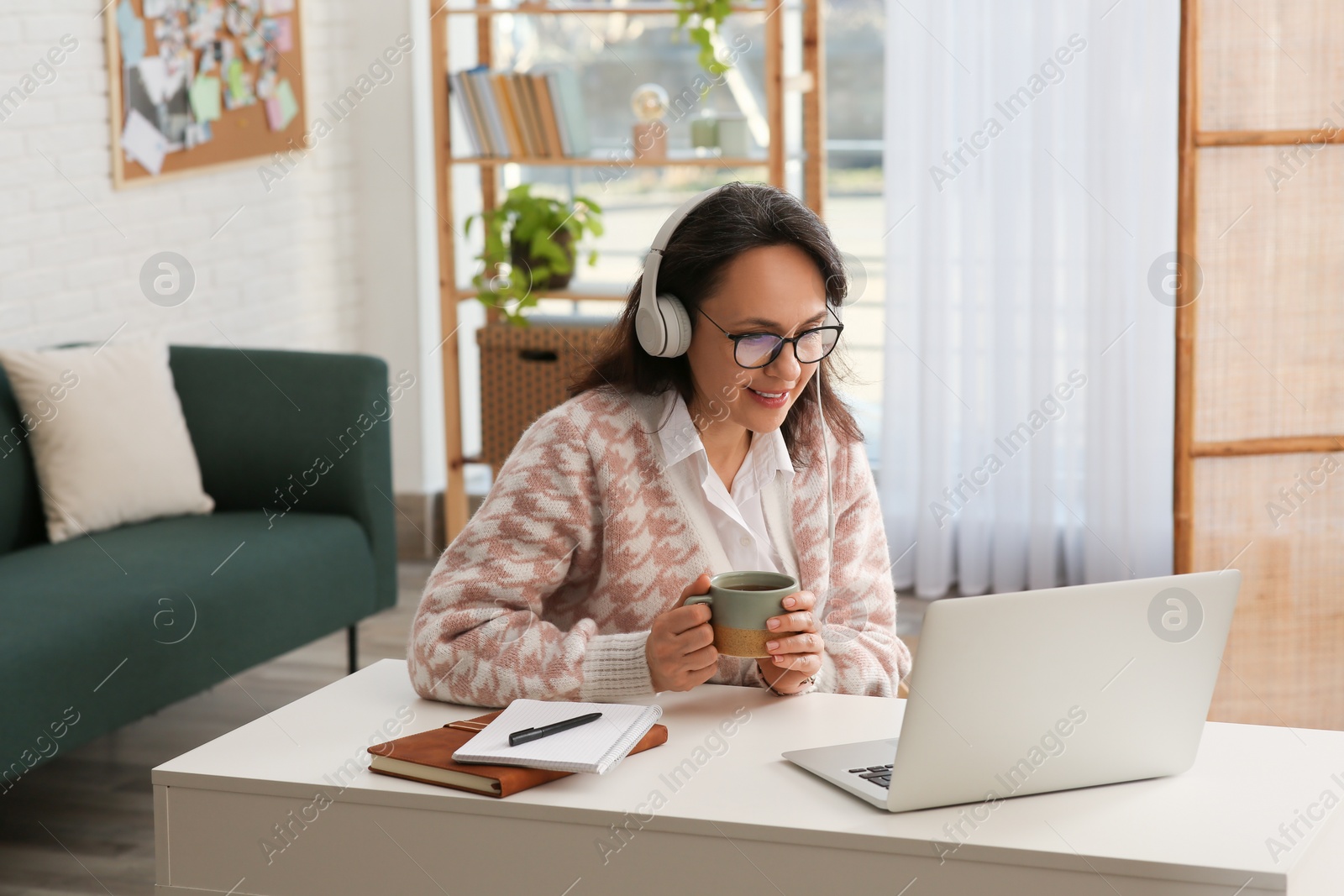 Photo of Woman with modern laptop and headphones drinking tea while learning at table indoors